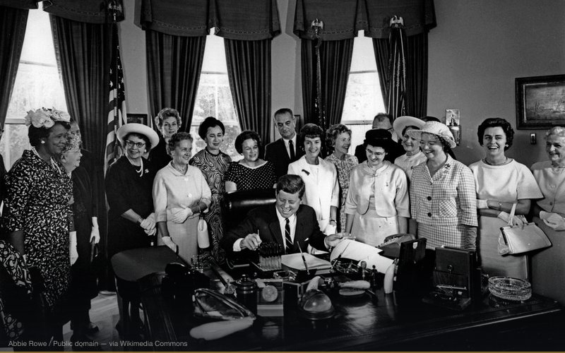 President John F. Kennedy (laughing) signs the Equal Pay Act in the Oval Office of the White House, Washington, D.C. Standing (L-R): President of the National Council of Negro Women (NCNW), Dorothy Height; former Director of the Women’s Bureau in the Department of Labor, Mary Anderson; Representative Elizabeth Kee (West Virginia); unidentified woman in back; Representative Edith Green (Oregon); Representative Edna Kelly (New York); Representative Catherine May (Washington); Vice President Lyndon B. Johnson; Director of United Automobile Workers (UAW) Women’s Department, Caroline Davis; Senator Maurine Neuberger (Oregon); President of the National Federation of Business and Professional Women’s Clubs (NFBPWC), Dr. Minnie Miles; Representative Leonor K. Sullivan (Missouri); Executive Director of the National Council of Catholic Women (NCCW), Margaret Mealey; Representative Martha W. Griffiths (Michigan); Representative Julia Butler Hansen (Washington). Also listed in the President’s schedule is Director of Legislation for the General Federation of Women’s Clubs (GFWC), Mary M. Chittenden. — related to Bay of Pigs — CIA Betrayal of Kennedy