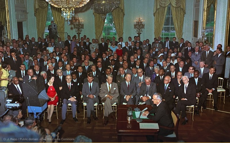 President Lyndon B. Johnson at the signing the 1964 Civil Rights Act. White House East Room. People watching include Attorney General Robert Kennedy, Senator Hubert Humphrey, First Lady "Lady Bird" Johnson, Rev. Martin Luther King, Jr., F.B.I. Director J. Edgar Hoover, Speaker of the House John McCormack. Television cameras are broadcasting the ceremony. — related to J. Edgar Hoover Murdered for His Files