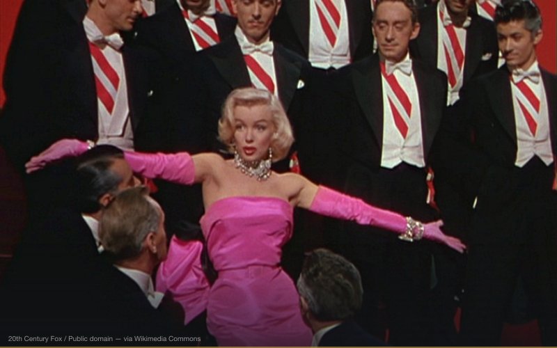 Gentlemen Prefer Blondes, the 1953 20th Century Fox movie is a musical comedy/romance directed by Howard Hawks starring Jane Russell and Marilyn Monroe, with Charles Coburn, Elliott Reid, Tommy Noonan, Taylor Holmes, and Norma Varden. This image shows Monroe in a pink dress surrounded by men in formal attire. — related to Marilyn Monroe, JFK & State Secrets