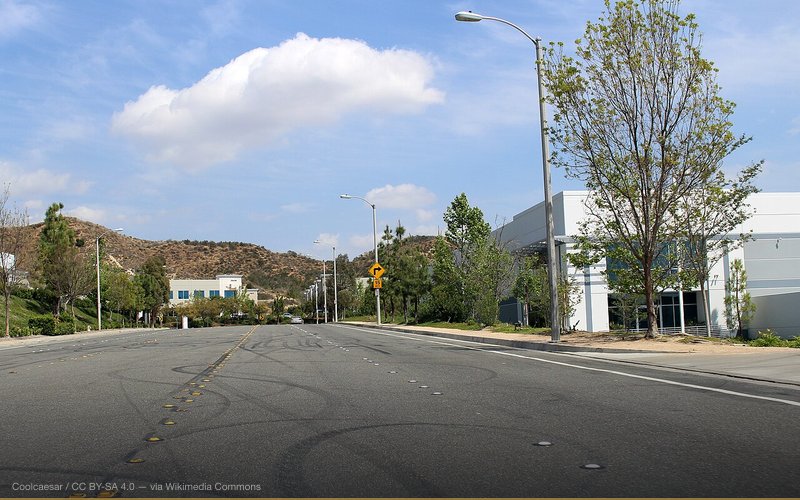 Hercules Street, facing eastbound, in the Rye Canyon Business Park in the Valencia neighborhood of Santa Clarita, California. According to media reports, the location on the right (just after the driveway) is reportedly where Paul Walker and Roger Rodas perished in a single-vehicle accident on November 30, 2013. The small objects on the ground barely visible to the right of the first light pole appear to be flowers left by fans. Photographed on April 5, 2015 by user Coolcaesar. — related to Paul Walker Was Murdered